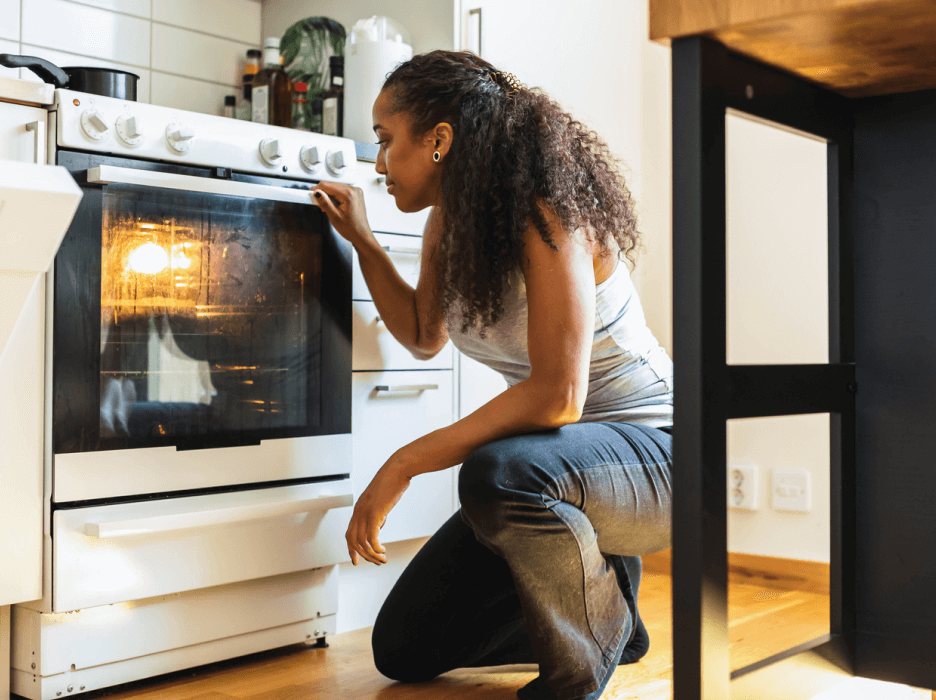 A woman is kneeling down, looking closely at her oven. 