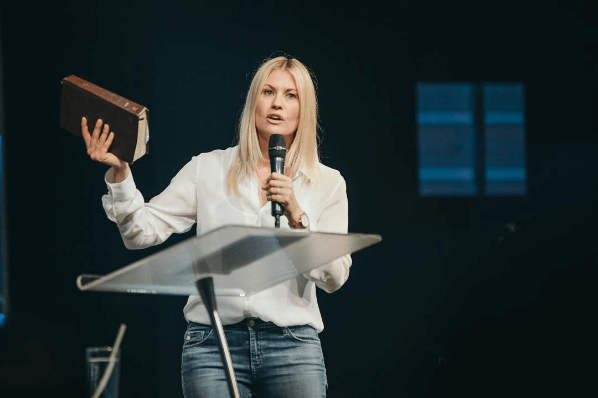 Holding a Bible and mic, Pastor Leanne preaches from behind a glass pulpit 