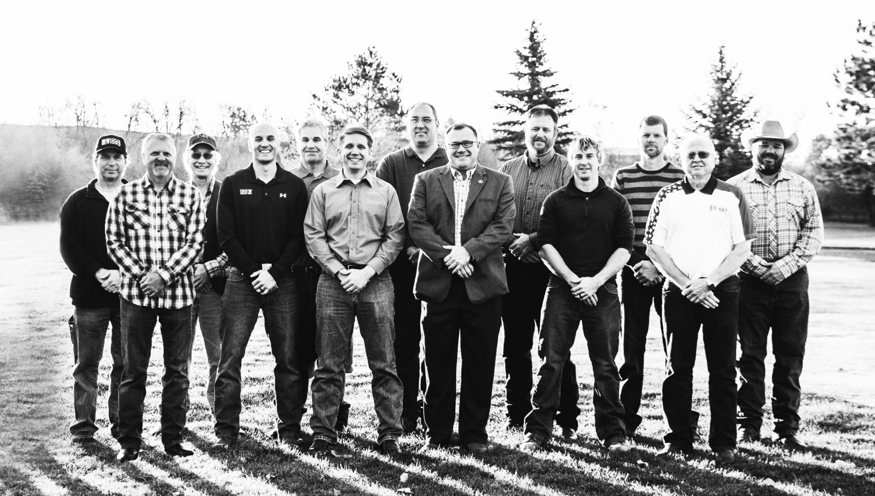 Black and white photo of 13 men wearing jeans and standing in a yard