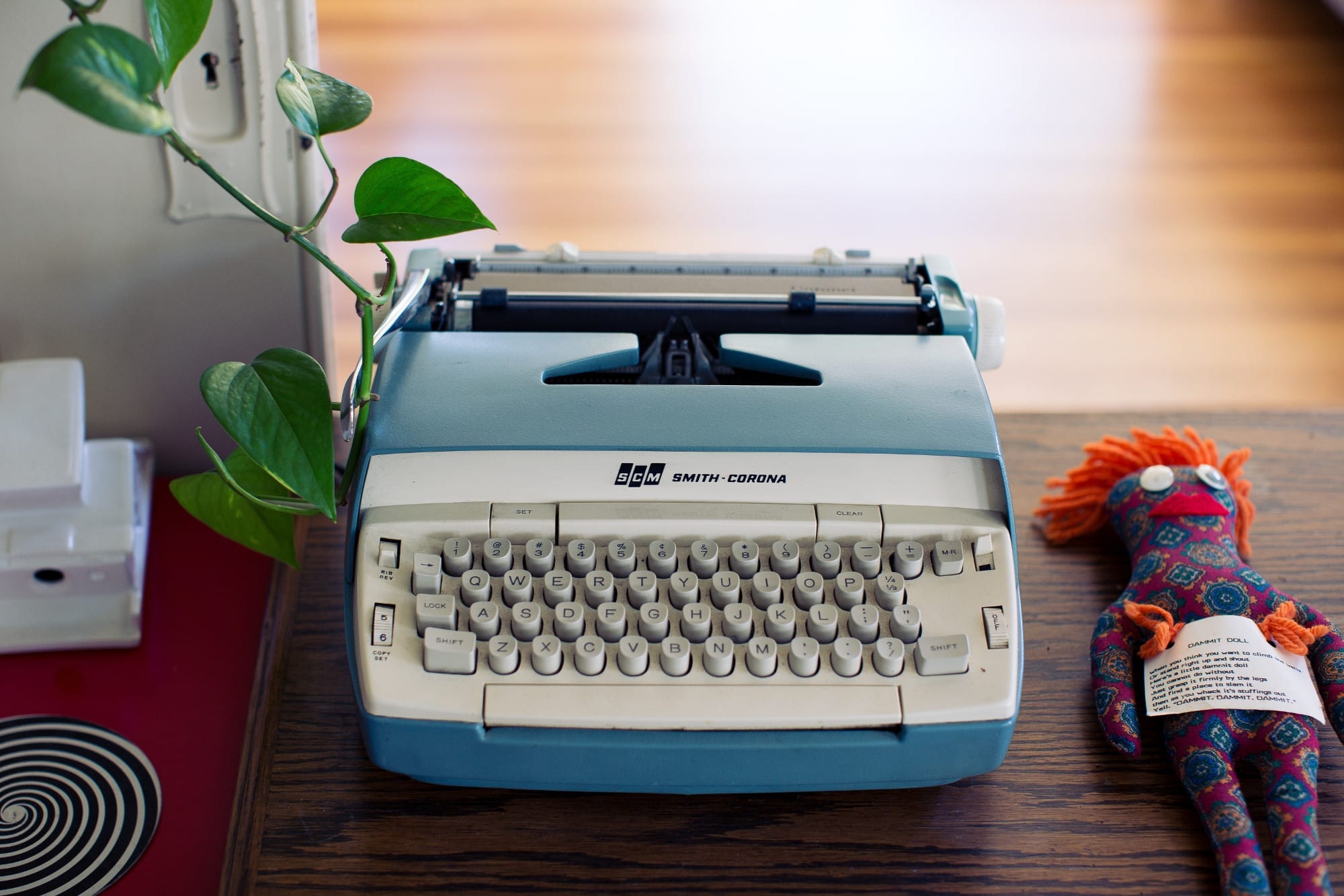 Baby blue vintage Smith-Corona typewriter on a desk next to a plant and a doll