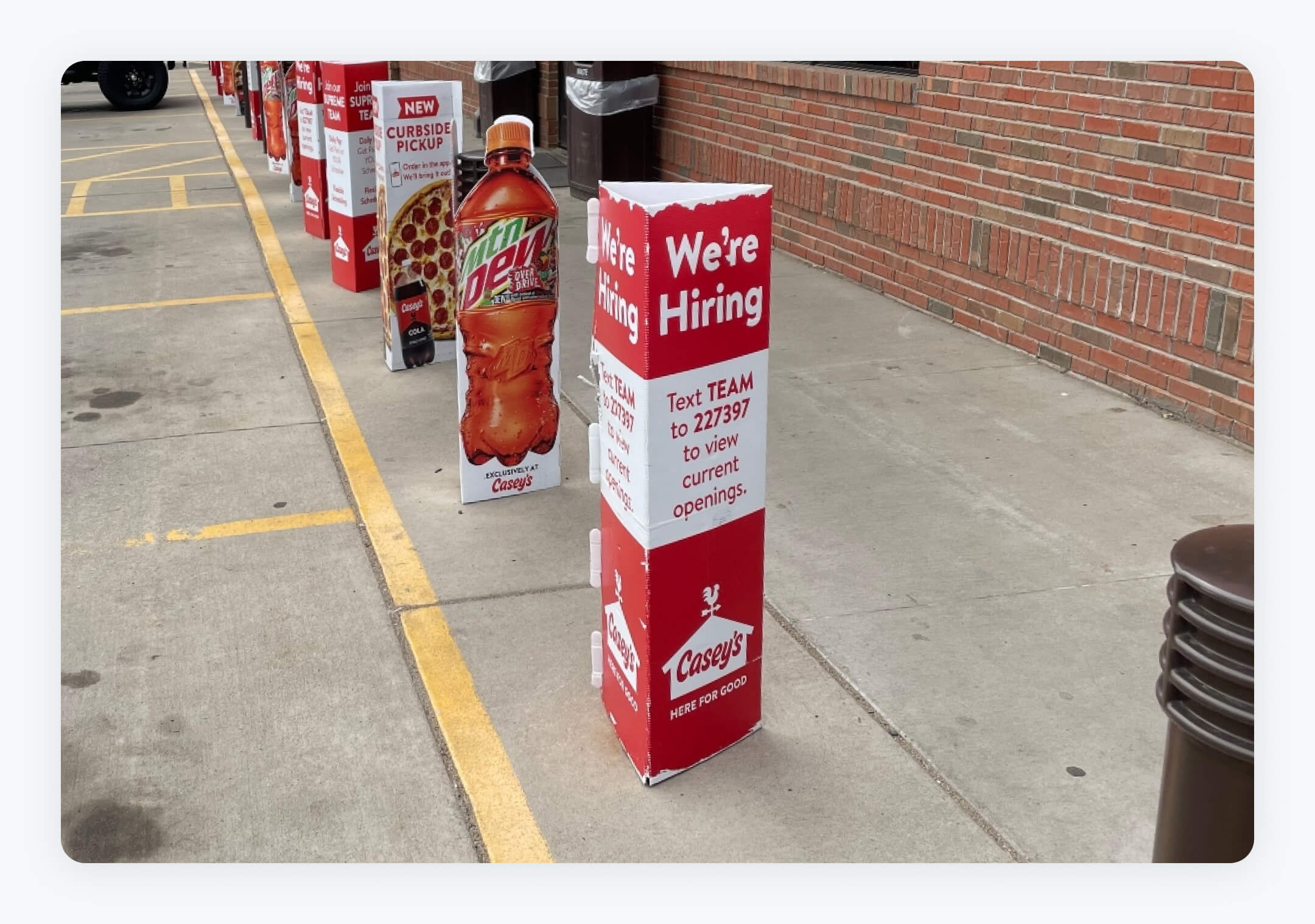 A row of vertical banner signs outside a brick building, with the closest sign prominently displaying 'We're Hiring' in large white letters on a red background. Below, it instructs to 'Text TEAM to 227397 to view current openings', aligning with the 'text to apply for job openings' initiative.